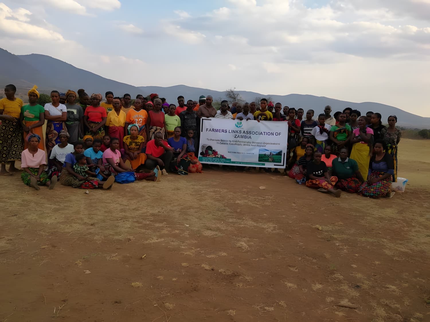 A large group of Farmers Links Association of Zambia members gathered outdoors in front of hills, holding a banner bearing the Association's name and mission statement.