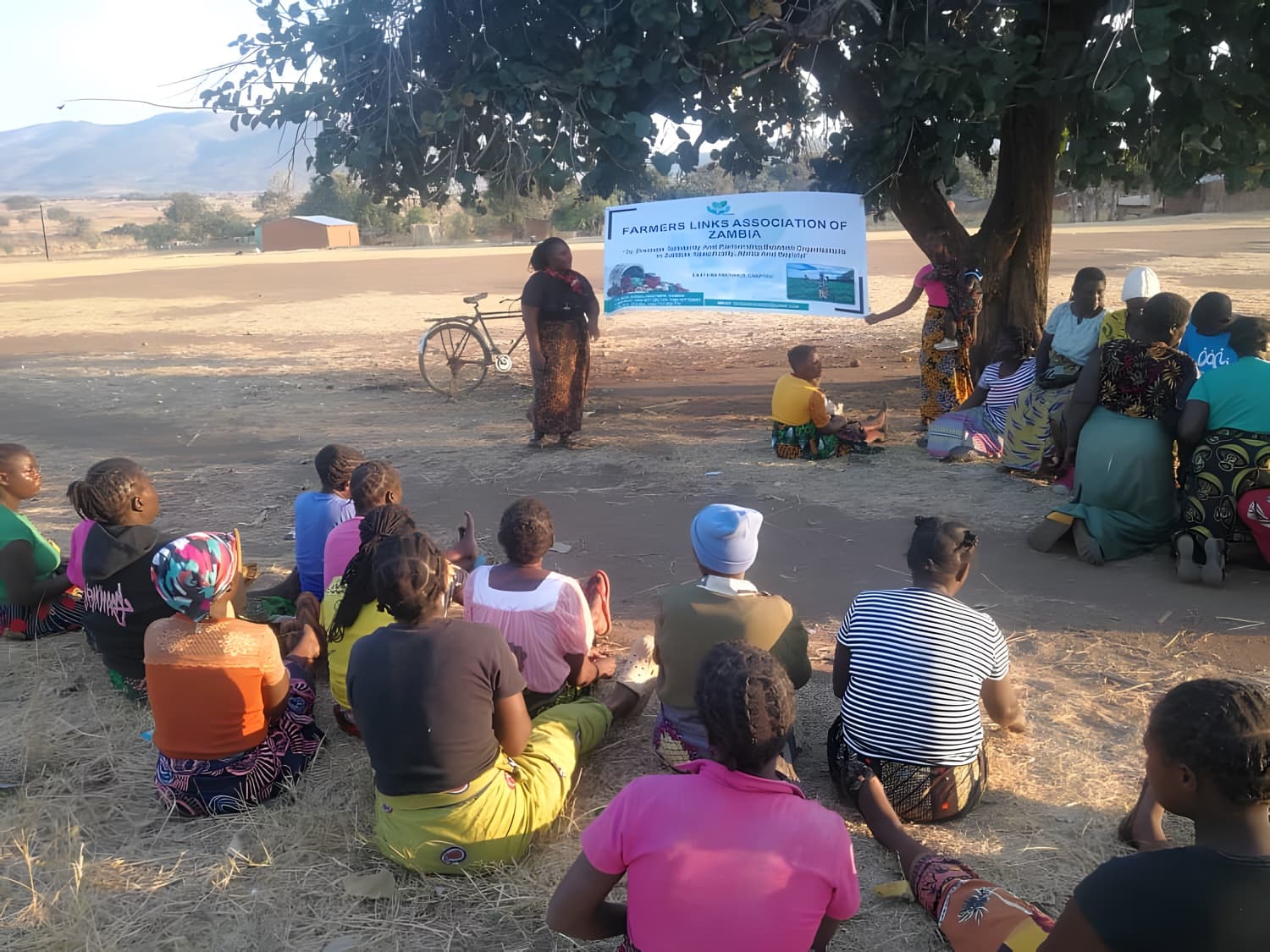 Women farmers seated under a tree at a Farmers Links training session as a facilitator addresses them, with hills behind.
